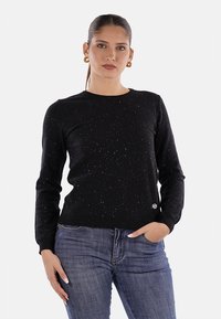 Donna con capelli legati, che indossa orecchini a cerchio dorati, un maglione a maniche lunghe speckled nero e jeans blu, in piedi con una mano in tasca.