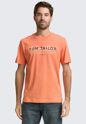 Mann trägt ein korallenoranges T-Shirt mit kurzen Ärmeln und dem Schriftzug "Tom Tailor" sowie blaue Jeans, steht vor einem schlichten, hellen Hintergrund.