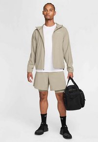 Man wearing beige jacket, shorts, white t-shirt, black sneakers and socks, holding a black gym bag against white background.