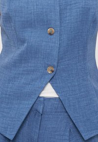 Veste texturée bleue avec deux boutons marron portée sur un top blanc, dévoilant un pantalon bleu assorti sous l’ouverture de la veste.