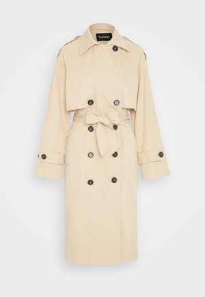 Cappotto beige con design corto, bottoni doppi neri, cintura da annodare e spalline; texture del tessuto liscia.