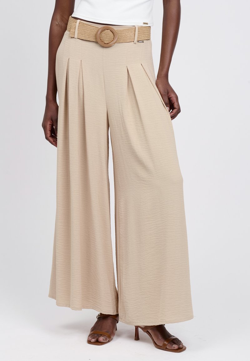 Pantalones beige de pierna ancha con pliegues, tejido texturizado y un cinturón tejido con hebilla redonda. Sandalias marrones de tacón completan el conjunto.