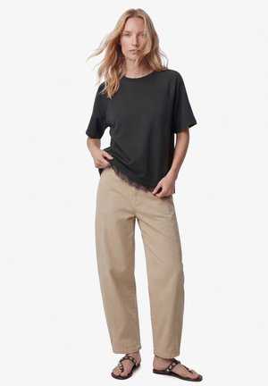 Ung kvinde med blondt hår iført en sort T-shirt med korte ærmer, beige vidde bukser og sorte pyntede sandaler, står mod en hvid baggrund.