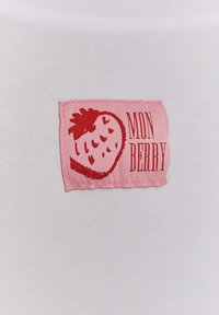 Bílá látka s růžovým obdélníkovým nášivkem, na kterém je design jahody a nápis "MON BERRY" v červené barvě. Nášivek je přišitý po okrajích.