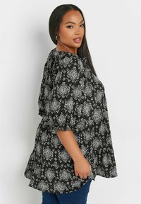 Czarna bluzka z białym, skomplikowanym kwiatowym wzorem, bufkami na rękawach i luźnym krojem. Stylizowana z niebieskimi dżinsami. Miękka, teksturowana tkanina.