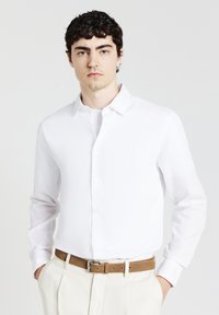 Camicia bianca a maniche lunghe con bottoni frontali, tessuto morbido e colletto classico. Indossata con pantaloni chiari e una cintura marrone.