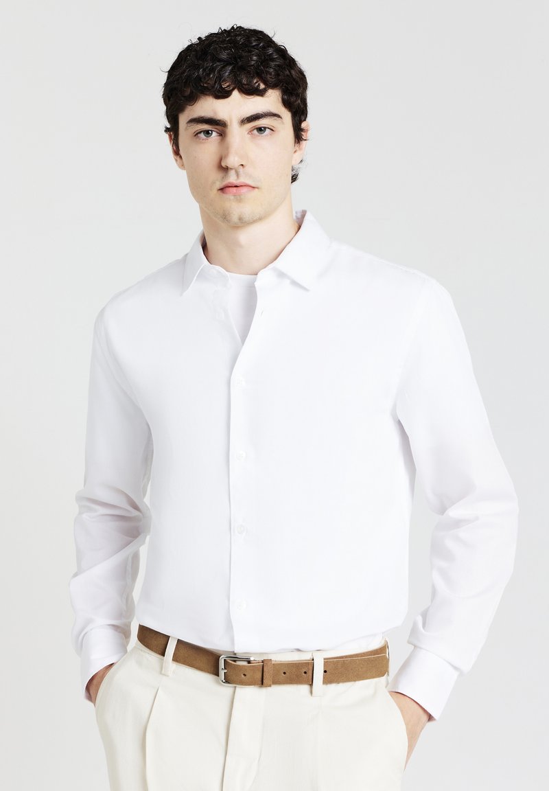 Camicia bianca a maniche lunghe con bottoni frontali, tessuto morbido e colletto classico. Indossata con pantaloni chiari e una cintura marrone.