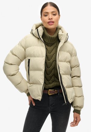Giacca puffer beige con cappuccio, dotata di tasche laterali con zip e polsini a costine. Indossata sopra un maglione verde a trecce e jeans scuri.