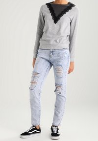 Sudadera gris con encaje negro en el escote, combinada con jeans claritos desgastados y zapatillas negras de corte bajo.