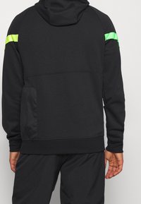 Haut à capuche noir avec un design épuré, des accents vert fluo sur les épaules, une poche kangourou et une capuche confortable. Texture lisse.