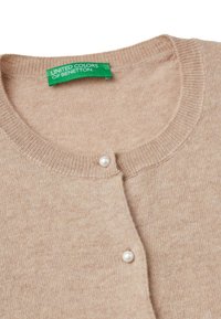 Cardigan en tricot beige avec un col rond, doté d'une fermeture par un seul bouton en perle et d'une étiquette verte indiquant "United Colors of Benetton."
