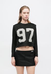 Schwarzes langärmliges Crop-Top aus Netzmaterial; mit weißem "97" Zahlenprint. Kombiniert mit einem hoch taillierten schwarzen Minirock mit Branding.