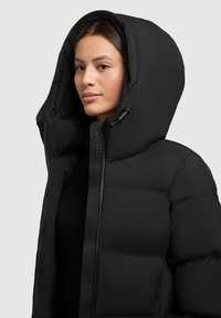 Chaqueta de plumas negra con alto cuello, capucha oversized y costuras prominentes, hecha de un tejido liso e insulated con cierre de cremallera.