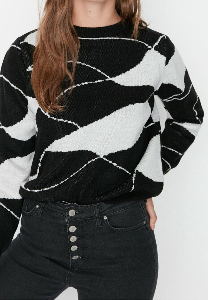 Femme portant un pull à motif abstrait noir et blanc et un jean noir taille haute avec cinq boutons visibles, main sur la hanche.