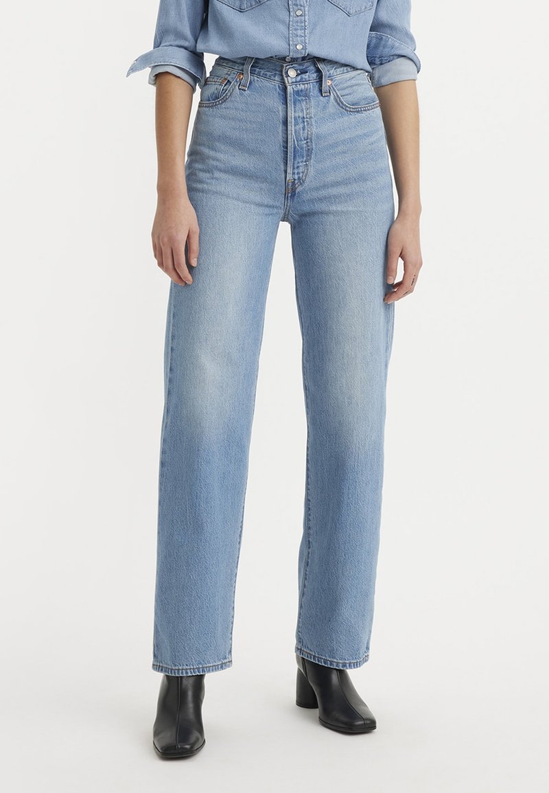 Femme portant un jean droit taille haute bleu clair, une chemise en denim retroussée et des bottines en cuir noir, debout devant un fond blanc.