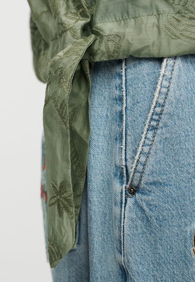 Πουκάμισο από σατέν ελιάς με κεντημένες λεπτομέρειες, δεμένο στη μέση, συνδυασμένο με ανοιχτό μπλε τζιν παντελόνι με φερμουάρ και κούμπωμα με κουμπί.