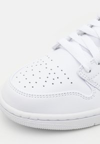 Gros plan sur l'avant d'une basket blanche avec de petits trous de ventilation, des lacets blancs et une couture détaillée sur une semelle texturée.