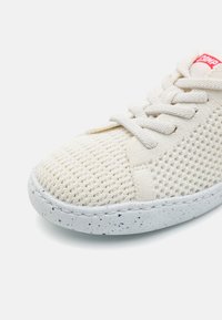 Sneaker with a cream mesh upper, textured fabric, and round toe. Features beige laces and a speckled white rubber sole. Red brand tag visible.