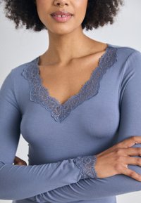 Femme avec les bras croisés portant un haut à manches longues lavande avec des bordures en dentelle au niveau de l'encolure et des poignets, affichant un piercing au septum et des lèvres naturelles.