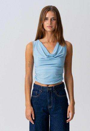 Mladá žena s rovnými hnedými vlasmi nosí svetlomodrý bezrukávový závesný top a tmavomodré rifle s vysokým pásom na jednoduchom pozadí.