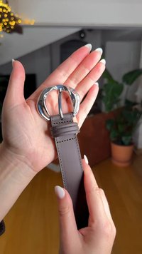 Ceinture en cuir marron avec une boucle argentée de forme sculpturale. La ceinture présente des détails de couture et est tenue dans une main sur un fond neutre.