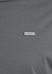 Grå, textured stoftop med et lille sølvmetal mærke, der læser "SMILODOX" og er fastgjort foran. Simpelt design, let materiale.