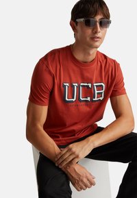 Červené bavlněné tričko s logem "UCB" v námořnické modré a bílé, krátké rukávy, kulatý výstřih, nošeno s černými kalhotami, model sedí s sepnutýma rukama.