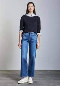 Vrouw binnenstaand, gekleed in een zwarte trui over een wit overhemd, blauwe high-waisted jeans en witte sneakers op een betonnen vloer.