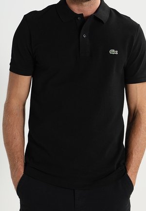 Mann trägt ein schwarzes Kurzarm-Poloshirt mit kleinem grünem Krokodil-Logo auf der linken Brust, Hände in den Taschen.