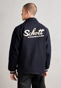 Jūras spēku vilnas jaka ar apkakli, aizmugurē ar izšūtu tekstu "Schott" un "NEW-YORK". Ribiņveida aproces ar baltām svītrām.