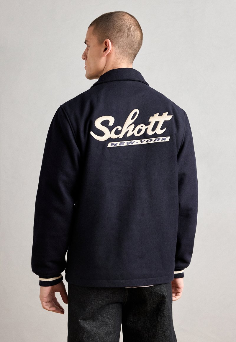 Jūras spēku vilnas jaka ar apkakli, aizmugurē ar izšūtu tekstu "Schott" un "NEW-YORK". Ribiņveida aproces ar baltām svītrām.