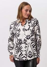 Woman wearing a white button-up shirt with black abstract floral patterns and black leather pants against a plain background.