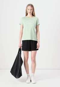 T-shirt en coton vert clair à manches courtes avec du texte blanc, associé à un short noir et un sac fourre-tout noir. Porté avec des baskets blanches.