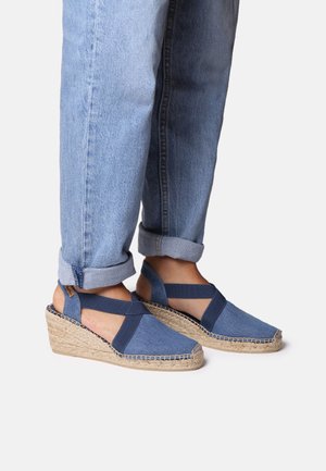 Modré džíny wedge espadrilky s designem překřížených popruhů a tkanou juteovou podrážkou. Mají strukturovaný svršek a prošívané detaily podél okrajů.