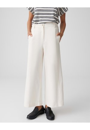 Person wearing wide-leg white pants, striped short-sleeve top, and black loafers, standing with hands in pockets on plain background.