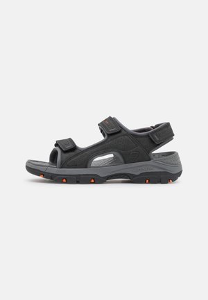 Sandalia outdoor Skechers negra y gris con tiras ajustables y detalles naranjas en la suela, mostrada en perfil lateral sobre fondo blanco.