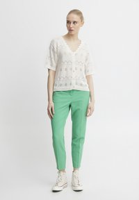 Weißer gestrickter Cardigan mit gewelltem Ausschnitt und Ausschnittmustern; kombiniert mit mintgrünen, maßgeschneiderten Hosen und weißen Sneakers.