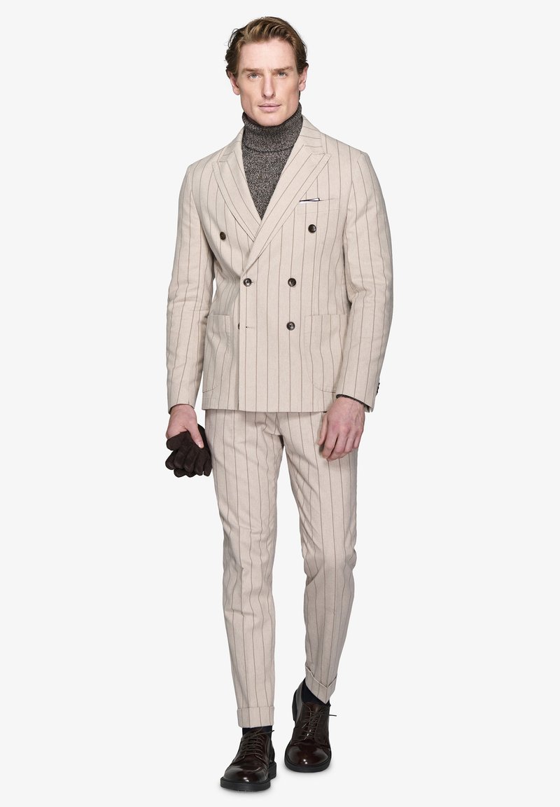 Abito beige a righe con doppiopetto, abbinato a un dolcevita grigio scuro a trama, guanti di pelle marroni e scarpe stringate scure. Vestibilità su misura.