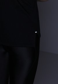 Czarna sportowa bluzka o perforowanej strukturze z srebrnym akcentem na dolnym końcu, w zestawieniu z błyszczącymi czarnymi legginsami dla uzyskania jednolitego wyglądu.