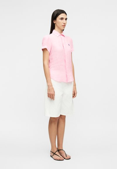 Femeie stând în picioare, cu fața spre înainte, purtând o cămașă roz deschis cu mâneci scurte și nasturi, pantaloni scurți albi până la genunchi și sandale negre.