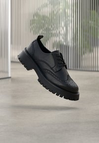 Μαύρο δερμάτινο παπούτσι brogue με διάτρητες λεπτομέρειες, χοντρή λαστιχένια σόλα και σχέδιο με κορδόνια. Ομαλή υφή με γυαλιστερό φινίρισμα.