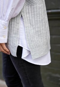 Pull en tricot gris à texture côtelée superposé sur une chemise blanche à col, avec des fentes sur les côtés et une finition lisse.