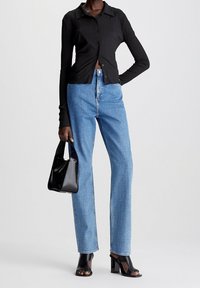 Camisa negra de botones con textura acanalada, vaqueros de tiro alto azul claro, tacones abiertos negros y bolso negro, todos sobre un fondo neutro.