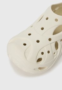 Chaussure beige clair à enfiler, fabriquée en matériau doux et texturé, dotée de multiples trous de ventilation et d'un design de semelle intérieure ergonomique.