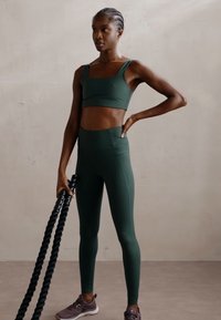 Ensemble de vêtements de sport en vert foncé, comprenant un soutien-gorge de sport ajusté et un legging taille haute, associés à des chaussures de sport foncées et une épaisse corde d'exercice.