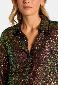 Camisa de lentejuelas con un patrón colorido y reflectante, con detalles de cuello y botones negros, que crea una apariencia texturizada y brillante.