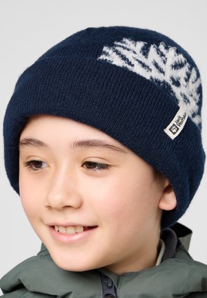 Gebreide marineblauwe beanie met een wit sneeuwvlokpatroon bovenop. Zachte textuur, opgerolde rand en een klein merklabel eraan bevestigd.