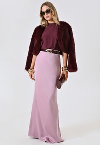 Burgunderfarbene Kunstfelljacke über einem kastanienbraunen Oberteil, pinke Satin-Rock, goldene Armreifen und eine kastanienbraune Clutch. Sonnenbrille rundet das Outfit ab.