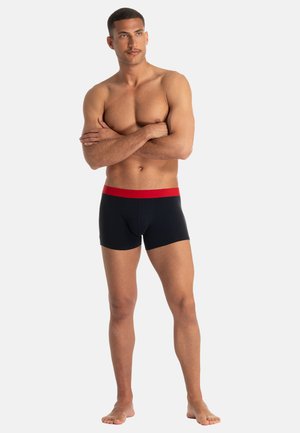 Sorte boxershorts med rød linning, lavet af glat stof, der sidder tæt. Modellen står barfodet med armene krydsede.