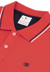 Rotes Poloshirt aus texturiertem Stoff mit einem marineblauen Kragen mit weißen Streifen, zwei Knöpfen und einem kleinen Logo auf der Brust.